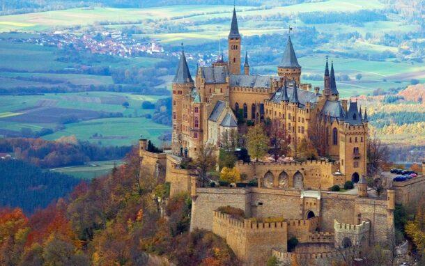 Die Burg Hohenzollern thront majestätisch auf einem bewaldeten Hügel, umgeben von herbstlich gefärbten Bäumen und weiten Feldern im Hintergrund.