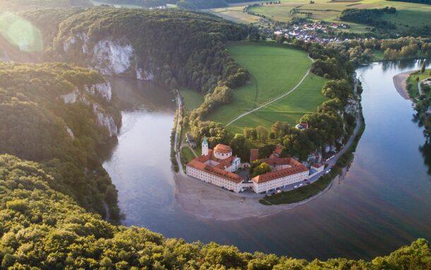 Luftaufnahme des Klosters Weltenburg an einer Flussschleife der Donau, umgeben von bewaldeten Felsen, Wiesen und kleinen Ortschaften.